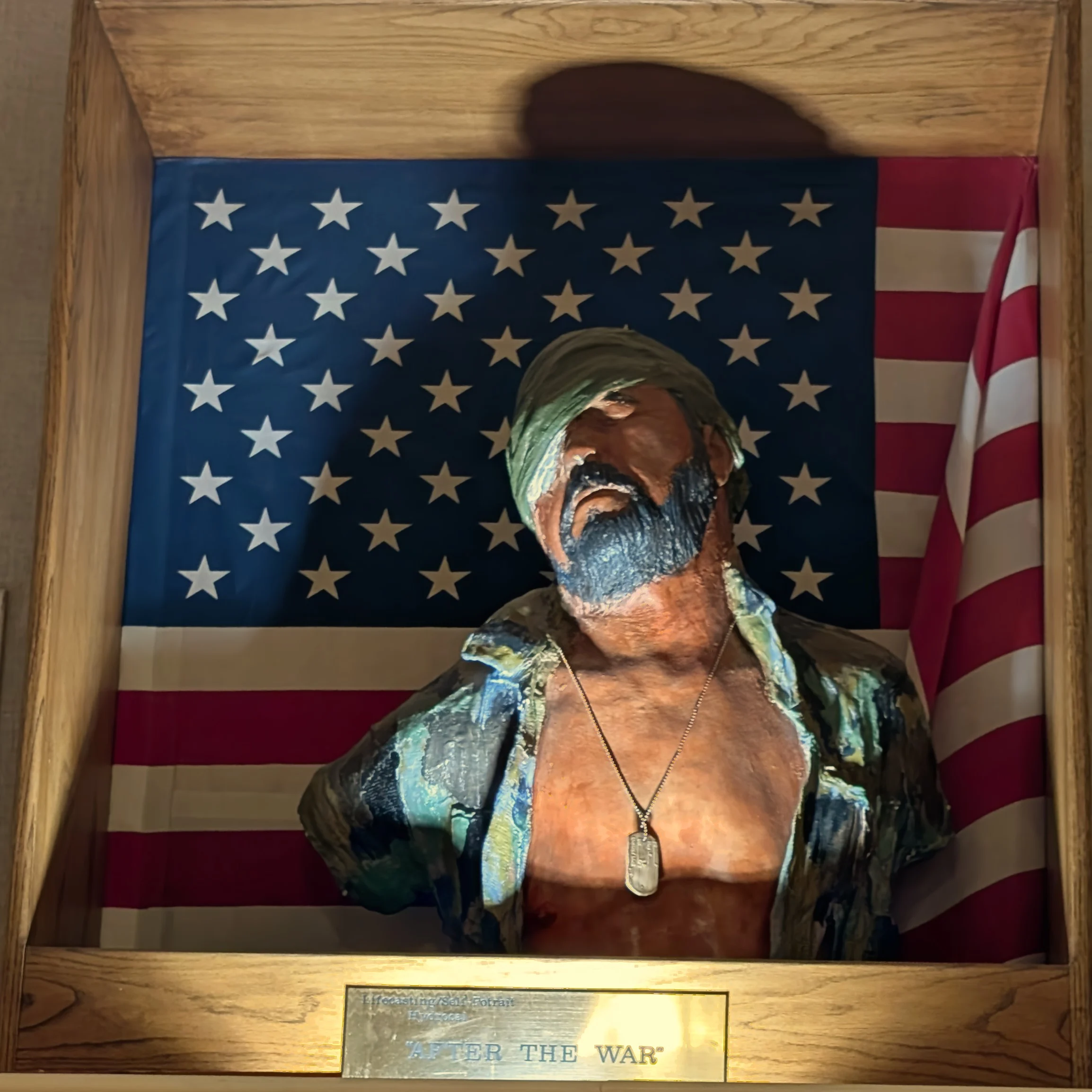 A life-cast bust of Rafael Gonzales displayed in a wooden case against an American flag. A green field bandage is wrapped across his eyes, his head is tilted back, and he wears an open shirt painted in camouflage blues and greens over a bare chest, with military dog tags around his neck. A brass plaque below reads: After the War.