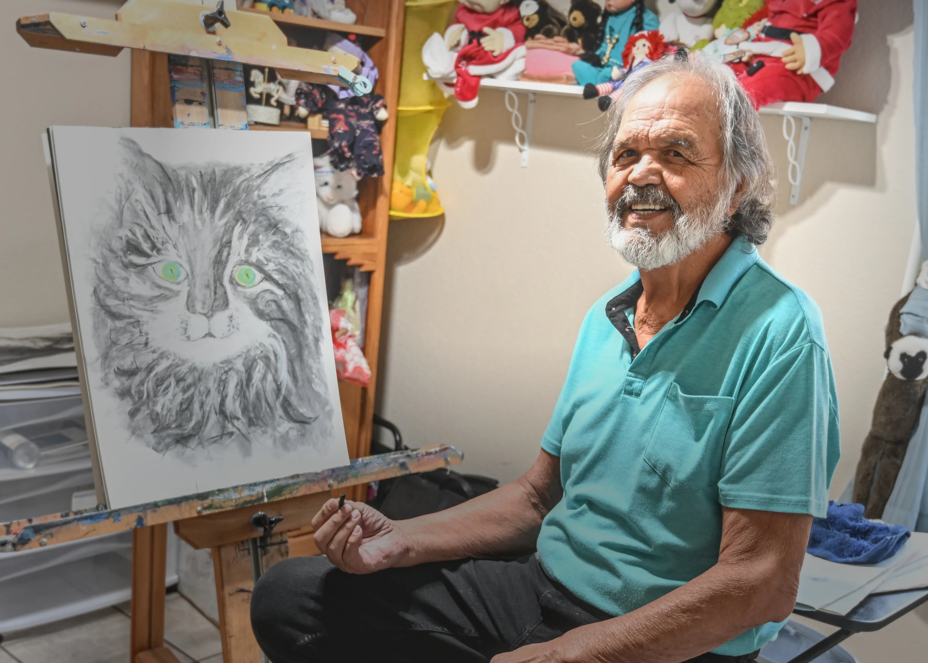 Rafael Gonzales at his easel in his studio, holding a piece of charcoal beside a charcoal drawing of a long-haired cat.
