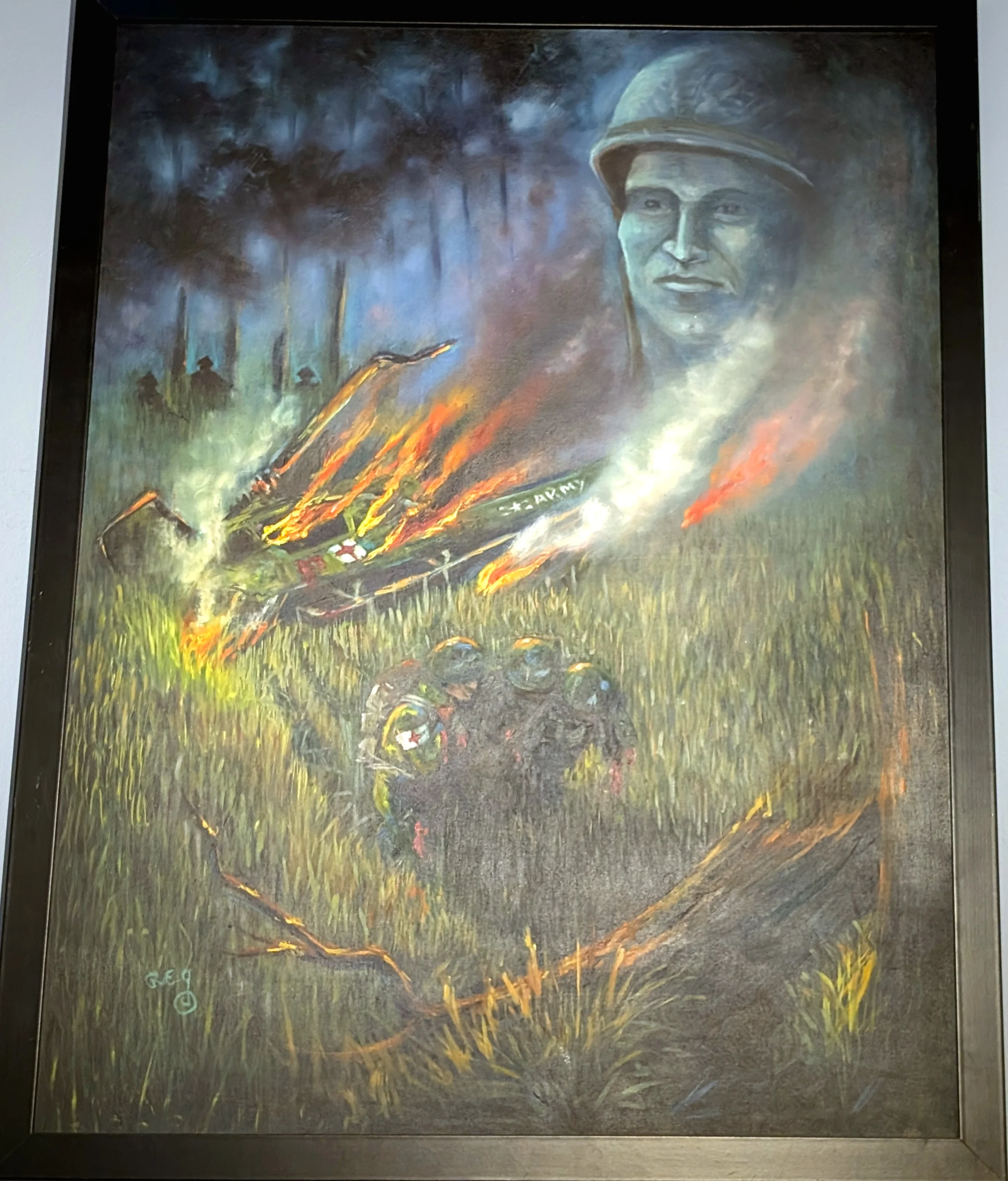 A painting of a U.S. Army helicopter burning in tall grass, flames and smoke streaming from it. In the foreground, figures lie low in the grass, one in a medic's helmet marked with a red cross. Shadowy shapes move among the trees in the dark background. In the upper right, the face of a soldier in a helmet rises above the scene like a spirit.
