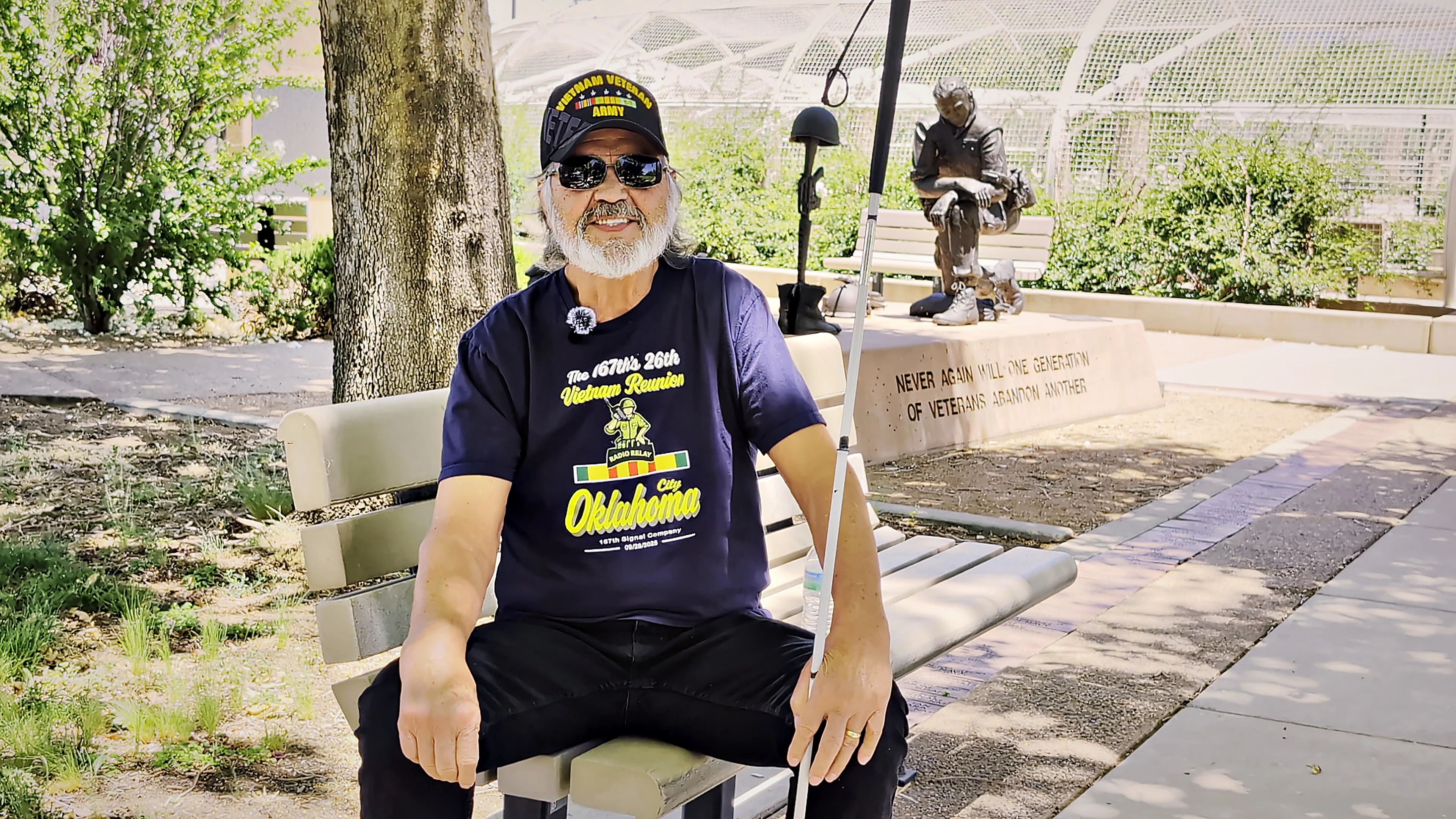 Rafael Gonzales, wearing a Vietnam Veteran Army cap and sunglasses, sits with his white cane on a bench at the Albuquerque Veterans Memorial. A bronze statue of a seated soldier stands behind him.